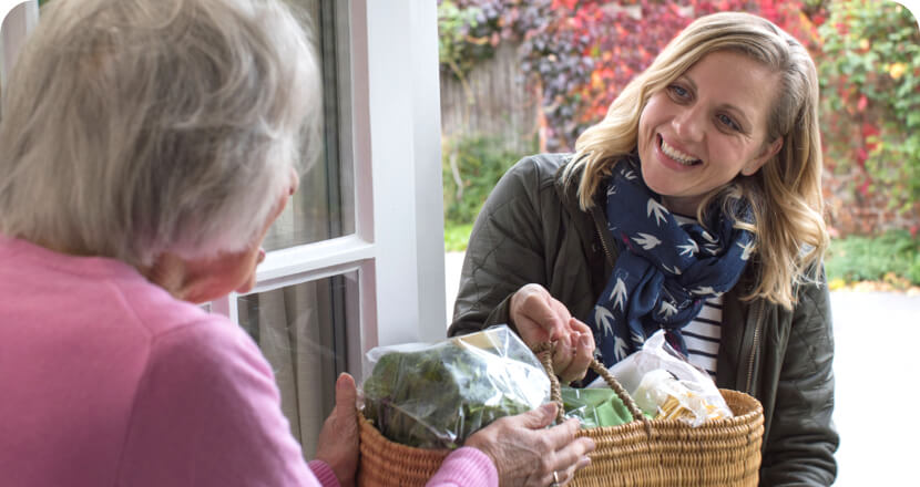 a woman smiles while handing over food basket to an elderly woman